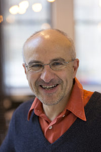 A smiling middle-aged man with glasses, a bald head, and a gray beard, wearing an orange collared shirt under a dark sweater, is standing indoors with blurred lights and windows in the background.