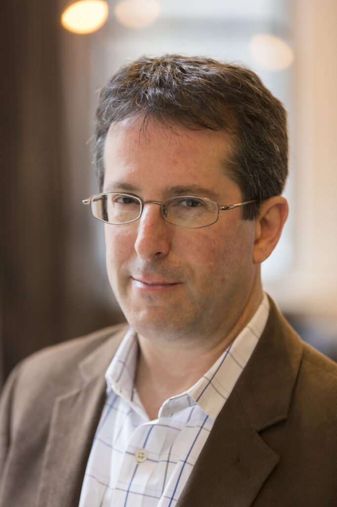 A middle-aged man with short brown hair, glasses, and a brown blazer over a white checkered shirt, looks at the camera with a neutral expression. The background is softly blurred.