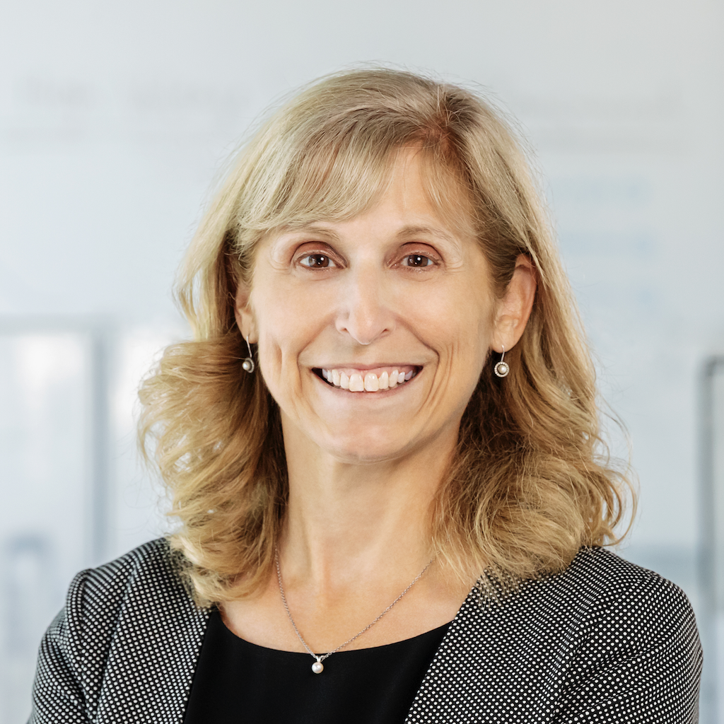 A woman with shoulder-length blonde hair, wearing a black top, a gray blazer, and pearl earrings, smiles in a bright office setting.