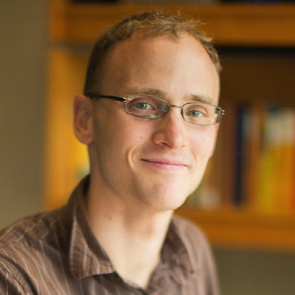 A person with short, light brown hair and glasses smiles softly, wearing a brown shirt. A blurred bookshelf is visible in the background.