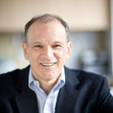 A middle-aged man in a dark suit jacket and light blue collared shirt smiles warmly at the camera indoors, with a blurred background.