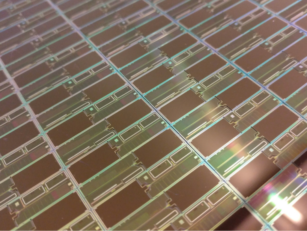 Close-up of a silicon wafer with multiple rectangular microchips, displaying intricate circuit patterns and reflecting light with a slight rainbow effect.
