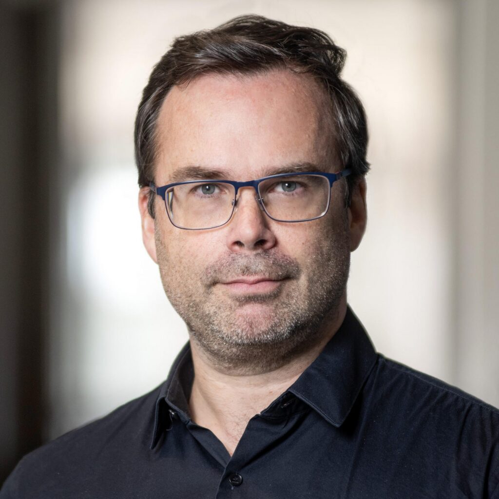 A man with short brown hair, glasses, and light facial hair, wearing a black collared shirt, looks at the camera with a neutral expression. The background is softly blurred.