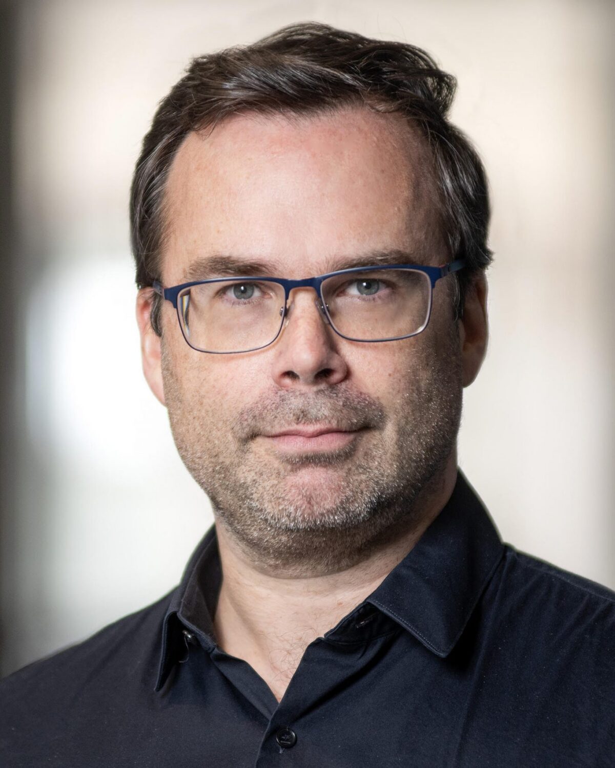 A man with short brown hair, glasses, and light facial hair, wearing a black collared shirt, looks at the camera with a neutral expression. The background is softly blurred.