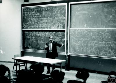 A professor stands and gestures in front of a large chalkboard filled with complex mathematical equations, addressing students seated in a lecture hall.