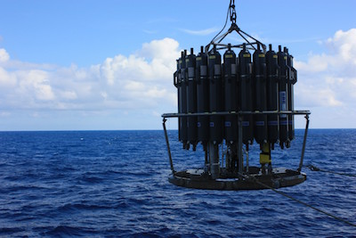A CTD rosette sampler is suspended above the ocean, ready to be lowered into the water for scientific research under a partly cloudy sky.