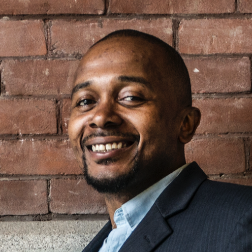 A man in a suit smiles while standing in front of a brick wall.