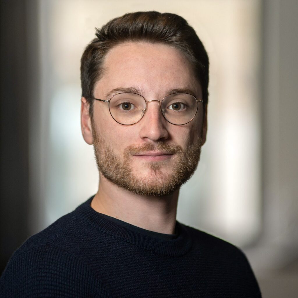 A man with glasses, short brown hair, and a trimmed beard wearing a dark sweater looks at the camera with a neutral expression. The background is softly blurred.