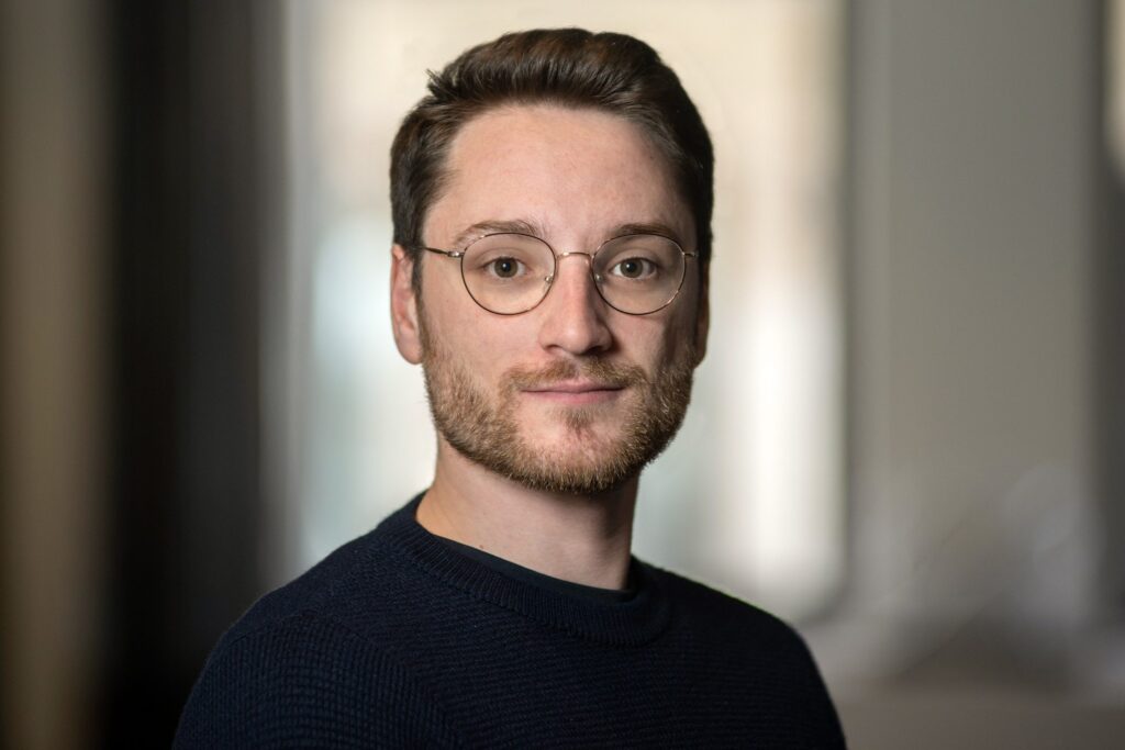 A man with glasses, short brown hair, and a trimmed beard wearing a dark sweater looks at the camera with a neutral expression. The background is softly blurred.