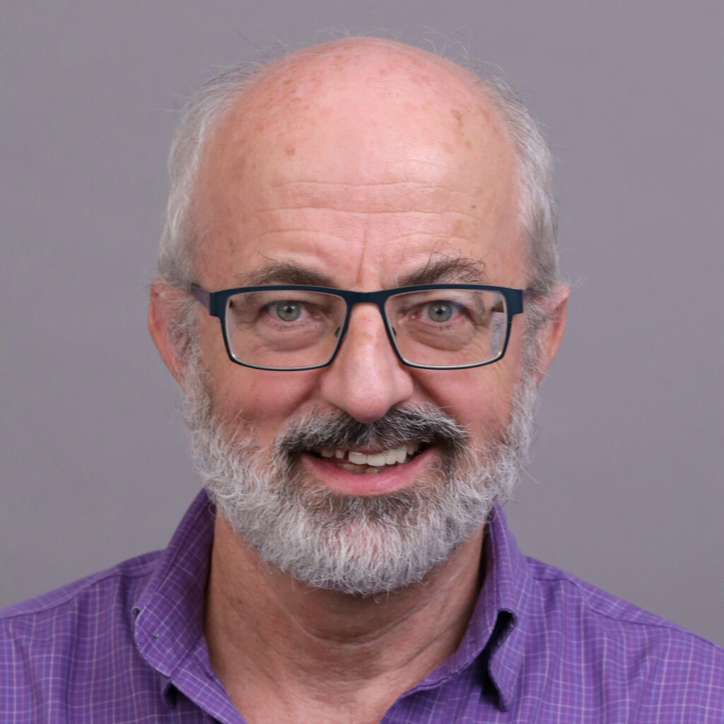 Smiling older man with glasses, gray hair, and a beard, wearing a purple collared shirt, in front of a plain gray background.