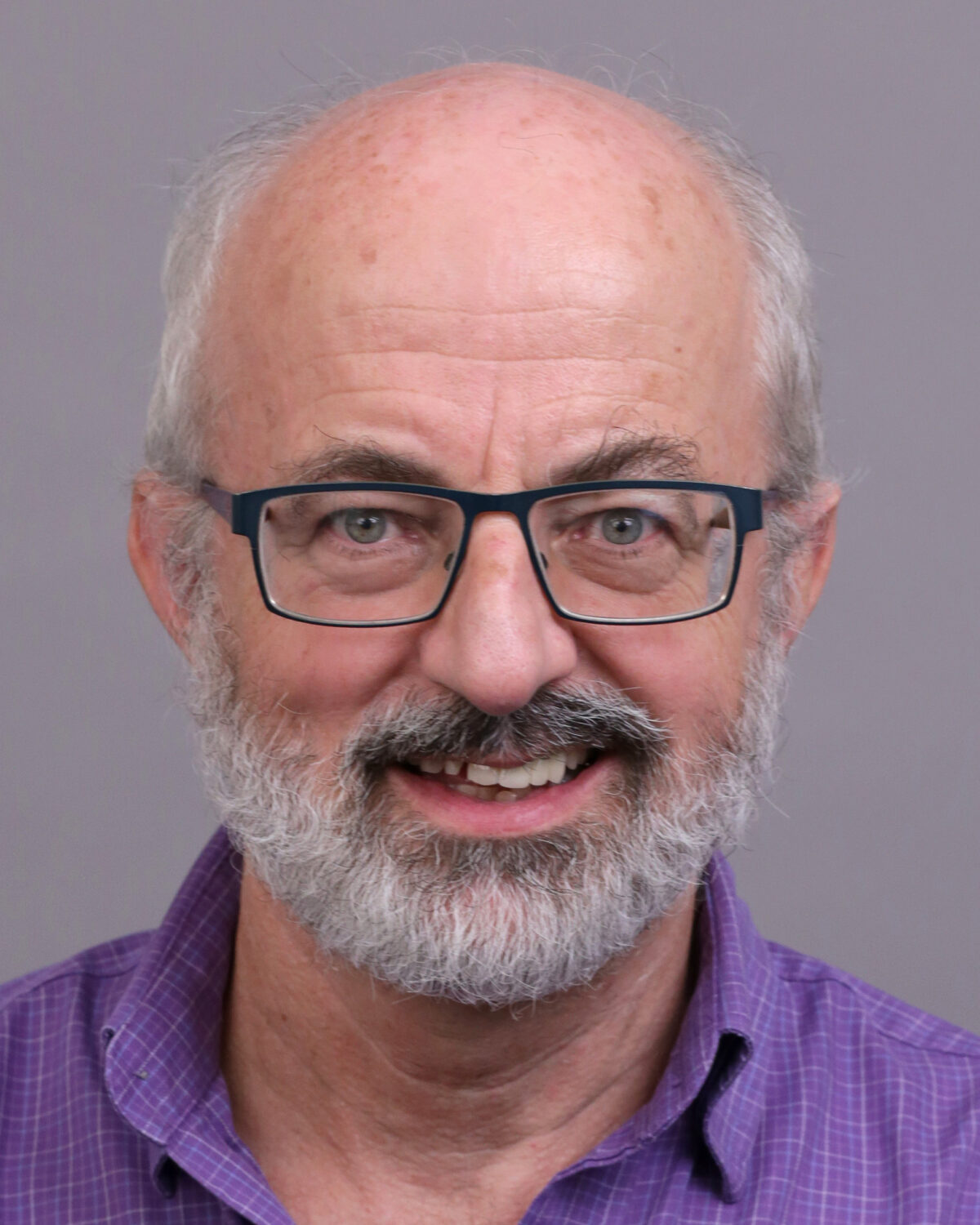 Smiling older man with glasses, gray hair, and a beard, wearing a purple collared shirt, in front of a plain gray background.