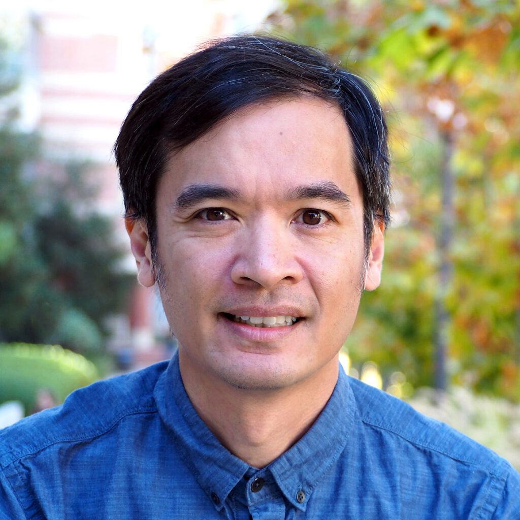 A man with short dark hair wearing a blue button-up shirt smiles outdoors with greenery and blurred trees in the background.