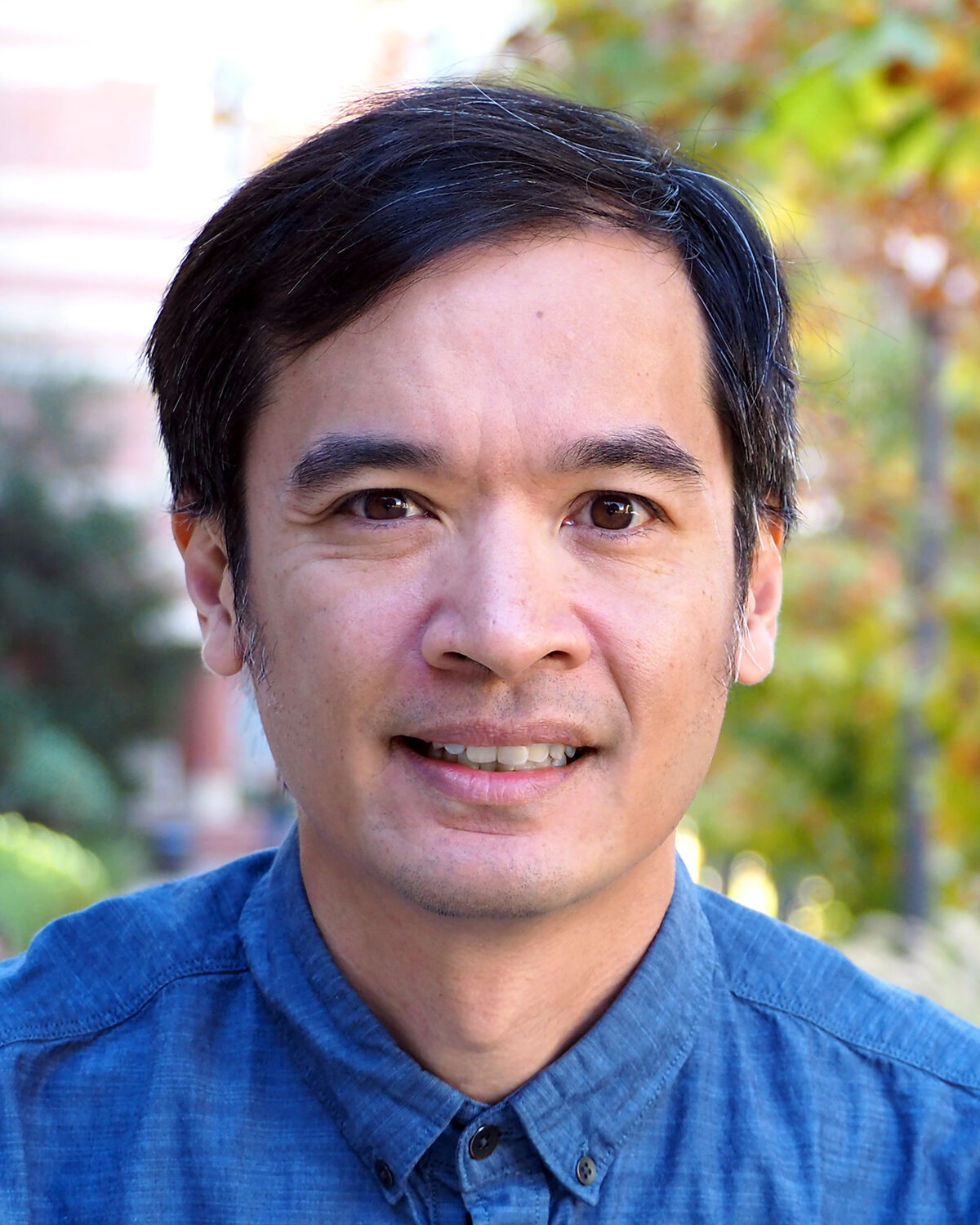 A man with short dark hair wearing a blue button-up shirt smiles outdoors with greenery and blurred trees in the background.