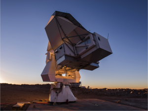 Large white radio telescope dish points upward at dusk, set against a clear sky and rocky landscape. The equipment is mounted on a sturdy base, silhouetted by the setting sun.