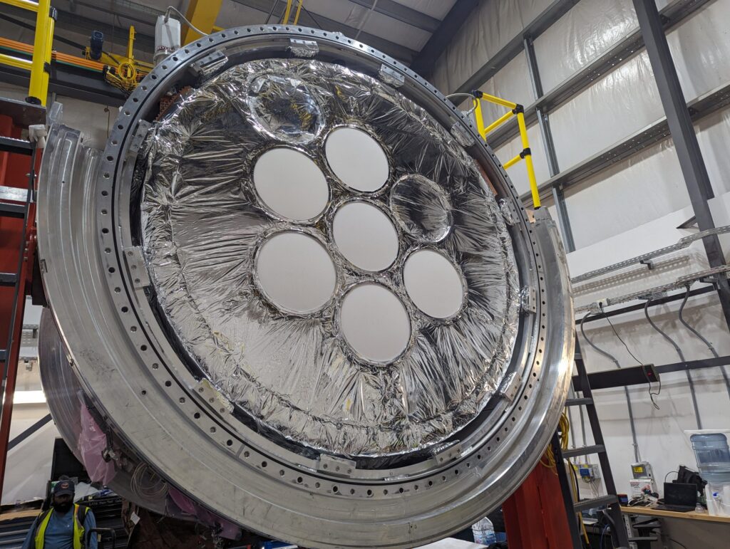 A large, circular metallic structure with seven white circular panels arranged in the center, surrounded by reflective foil, is seen inside an industrial facility with exposed beams and equipment.