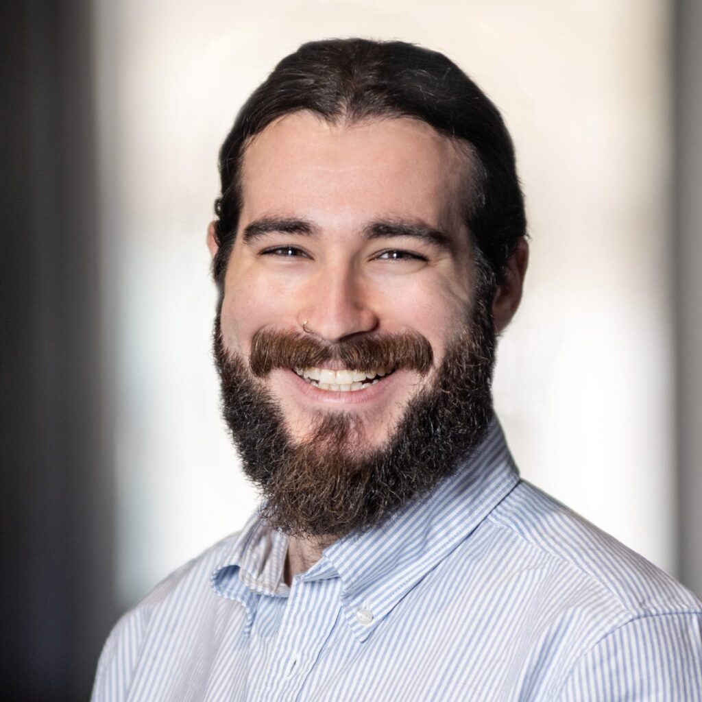 A man with long dark hair, a full beard, and a mustache smiles at the camera. He is wearing a light blue striped button-up shirt. The background is softly blurred.