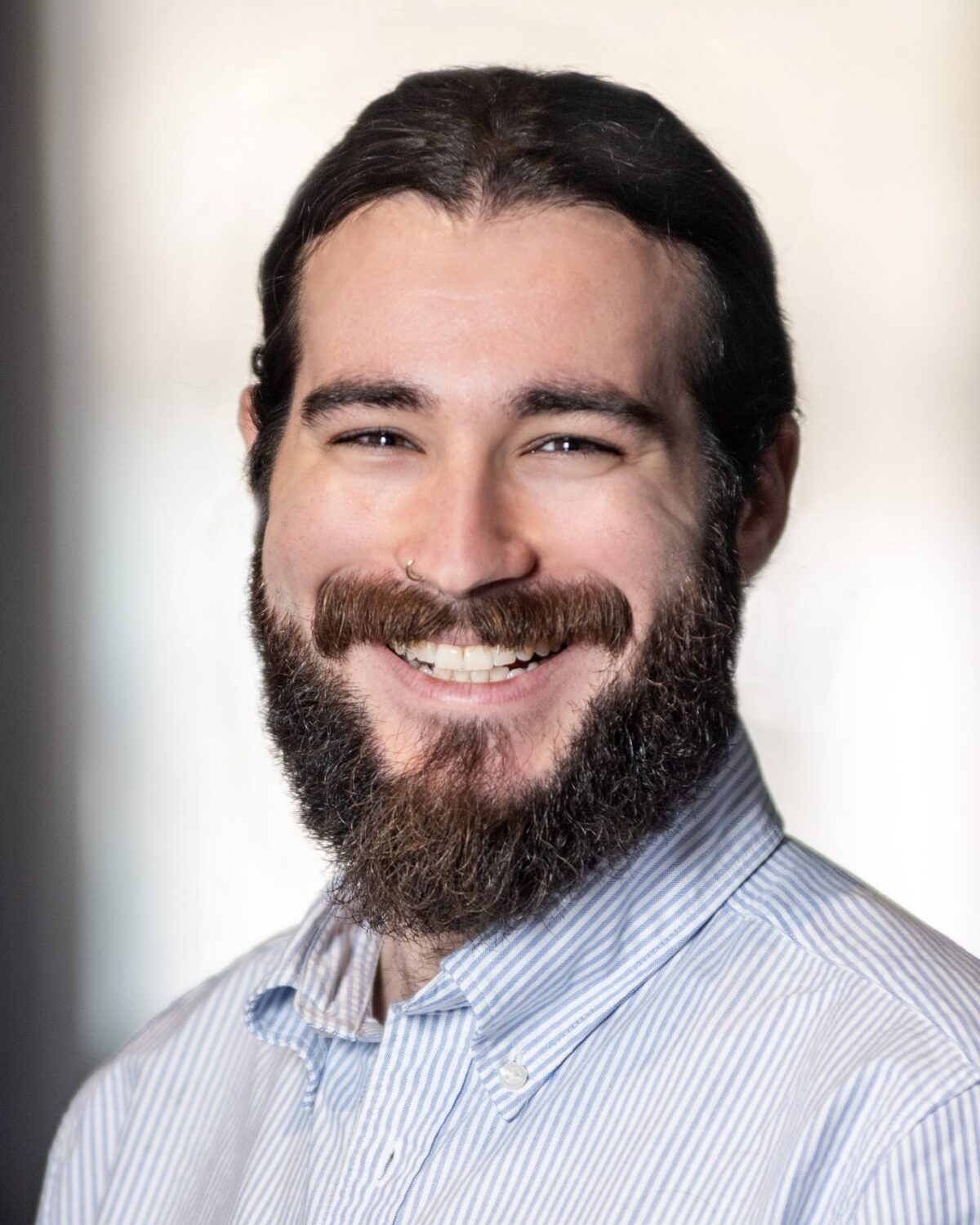 A man with long dark hair, a full beard, and a mustache smiles at the camera. He is wearing a light blue striped button-up shirt. The background is softly blurred.