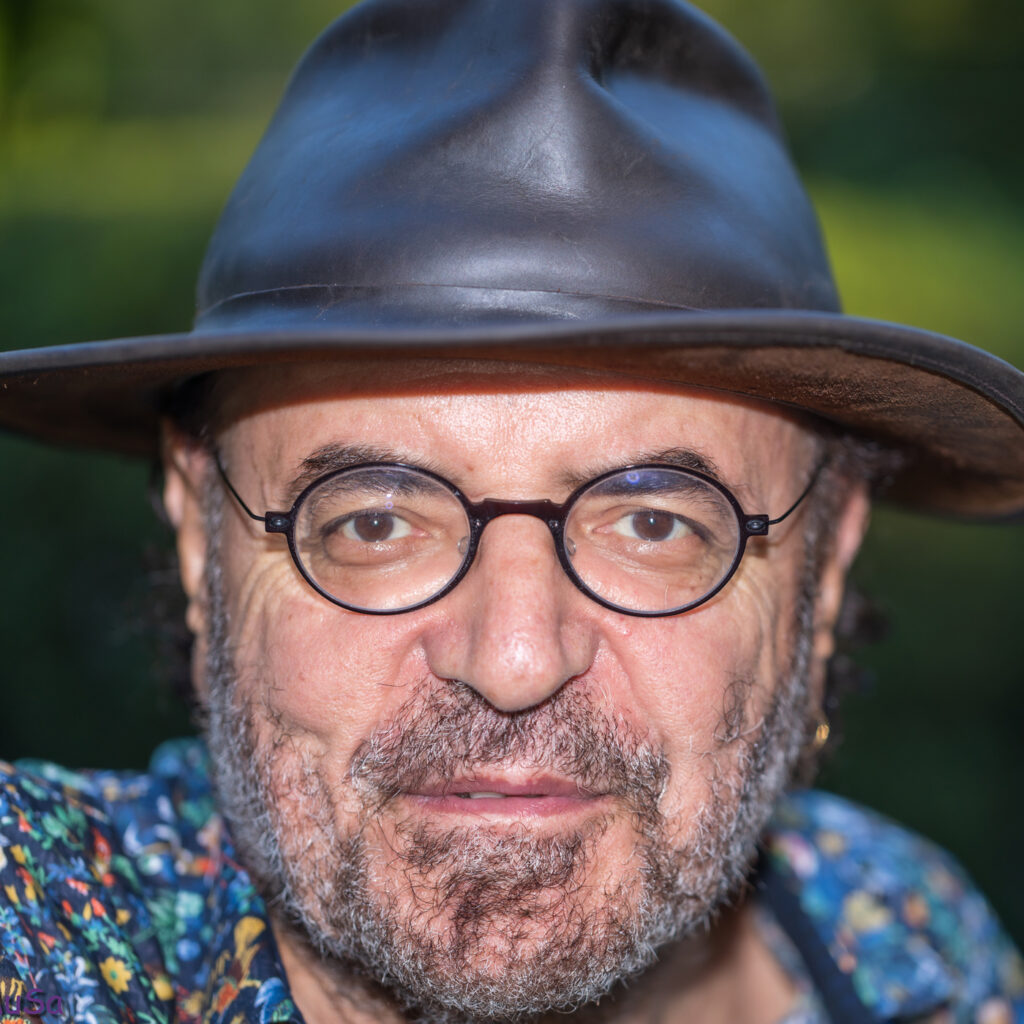 A man with a beard and mustache wears round glasses, a patterned shirt, and a wide-brimmed brown hat, standing outdoors with greenery blurred in the background.