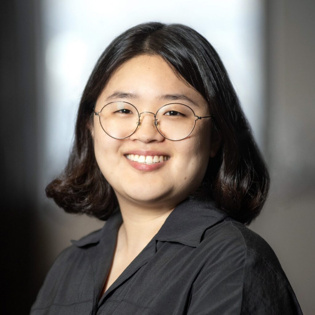 A person with short dark hair, glasses, and a black shirt smiles at the camera against a blurred indoor background.