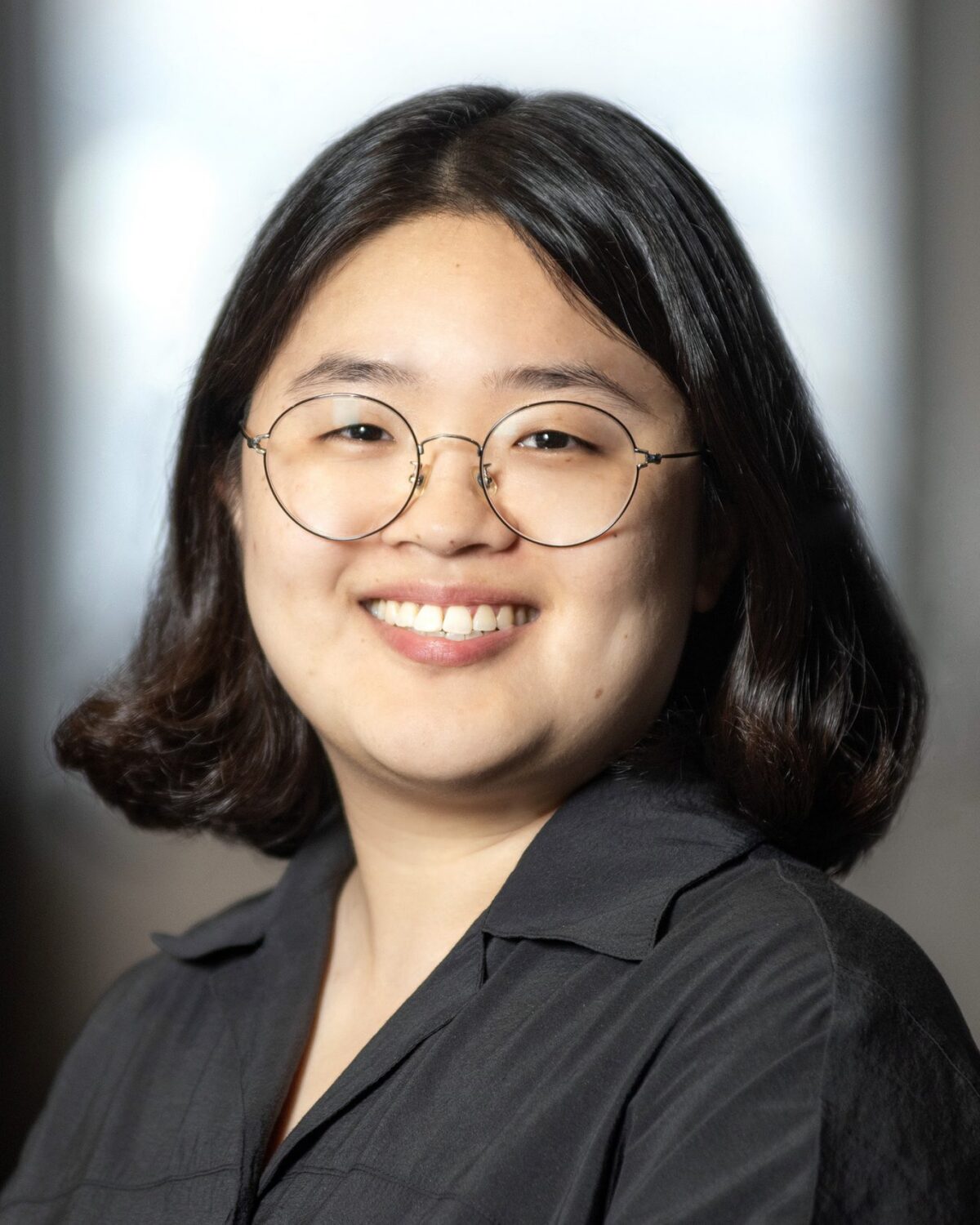 A person with short dark hair, glasses, and a black shirt smiles at the camera against a blurred indoor background.