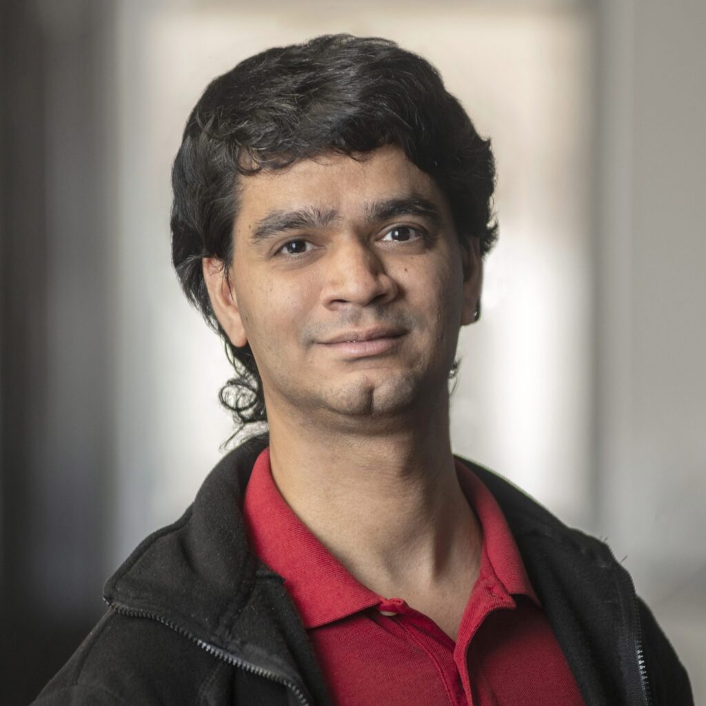 A person with dark hair is wearing a red collared shirt and a dark jacket, standing indoors in front of a blurred background, smiling softly at the camera.