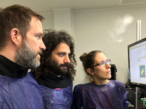 Three people in purple lab gowns closely examine data displayed on a computer monitor in a lab setting.