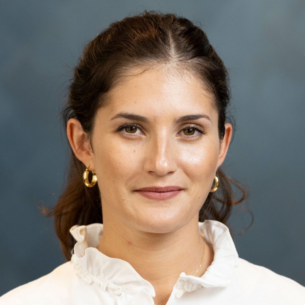 Woman with dark hair tied back, wearing gold hoop earrings and a white ruffled blouse, smiles softly against a plain blue-gray background.