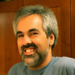A man with gray hair and a beard smiles warmly while wearing a blue shirt. The background appears to be a wooden surface, possibly a cabinet or panel.