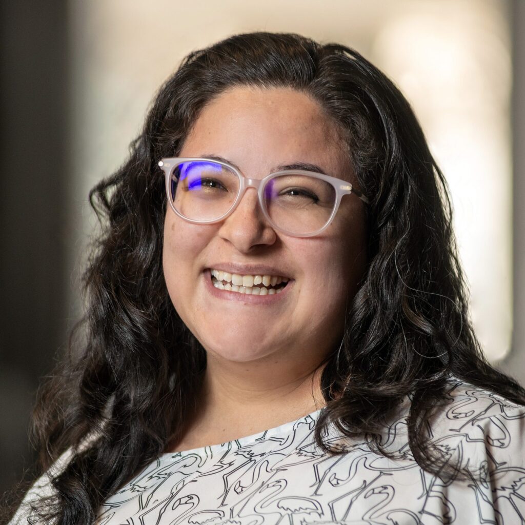 A woman with long dark hair and glasses smiles at the camera, wearing a patterned white blouse. The background is softly blurred.