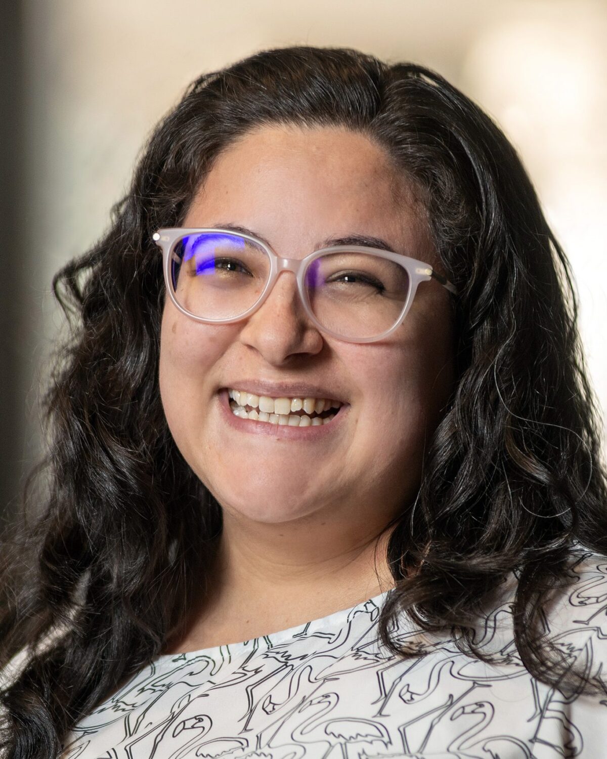 A woman with long dark hair and glasses smiles at the camera, wearing a patterned white blouse. The background is softly blurred.