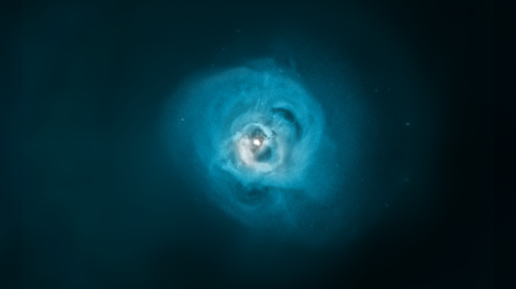 A bright blue nebula with swirling gas and dust clouds appears against a dark background, with a luminous center and faint specks of stars scattered throughout the image.