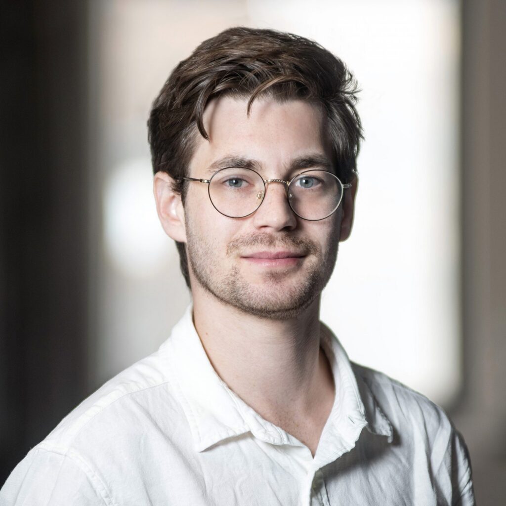 A young man with brown hair, glasses, and slight facial hair wears a white shirt and smiles softly, standing indoors with blurred background.