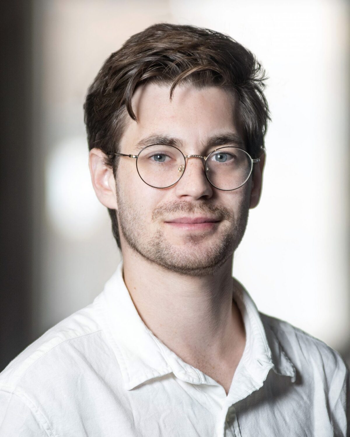 A young man with brown hair, glasses, and slight facial hair wears a white shirt and smiles softly, standing indoors with blurred background.