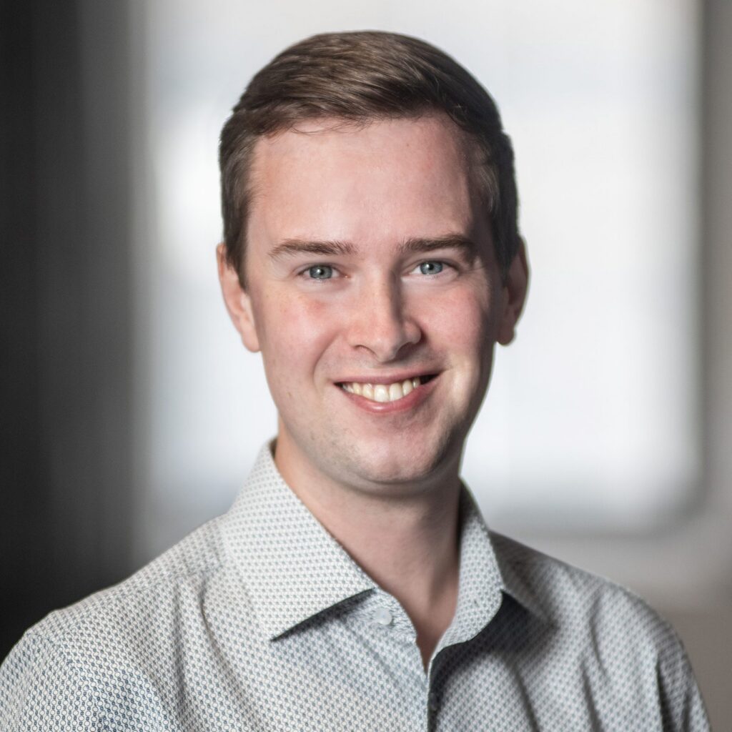 A young man with short brown hair smiles, wearing a light patterned shirt. The background is softly blurred with neutral tones, keeping the focus on his face.