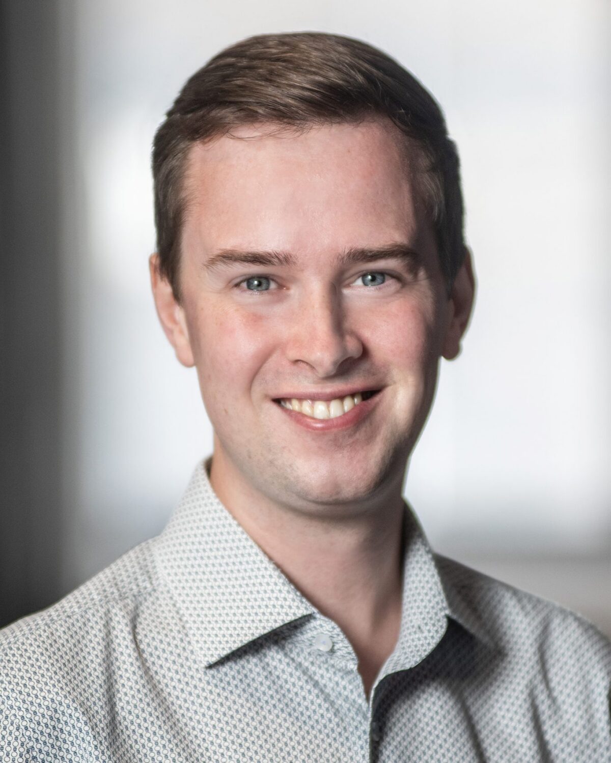A young man with short brown hair smiles, wearing a light patterned shirt. The background is softly blurred with neutral tones, keeping the focus on his face.