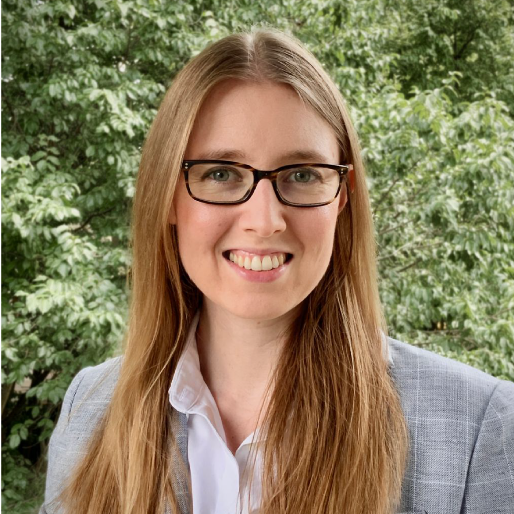 A woman with long blonde hair, wearing glasses, a light gray blazer, and a white shirt, smiles at the camera. Green trees fill the background.
