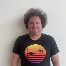A person with curly hair stands against a plain white wall, wearing a black T-shirt featuring a colorful sunset with silhouettes of spaceships.