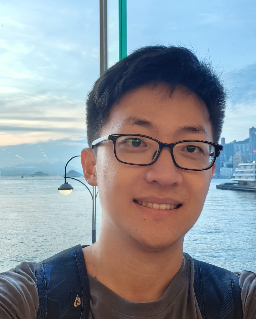 A person wearing glasses and a backpack smiles for a selfie near a waterfront with city buildings, a pier, and mountains visible in the background under a blue sky.