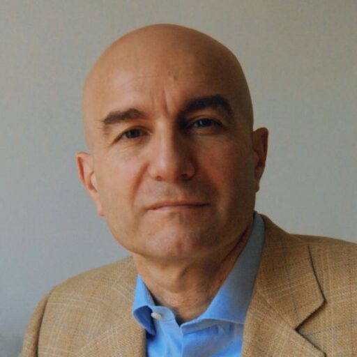 A bald man wearing a tan blazer and blue shirt looks at the camera with a neutral expression against a plain background.