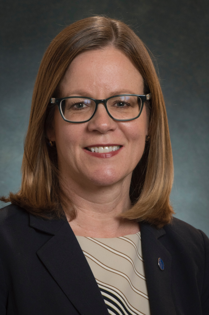 A woman with straight, shoulder-length brown hair and glasses smiles at the camera. She is wearing a dark blazer over a patterned blouse, with a blue pin on her lapel. The background is dark and plain.