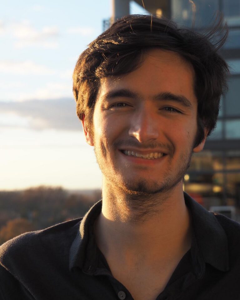 A young man with dark hair and a short beard smiles outdoors at sunset, with a blurred background of trees and a modern building.