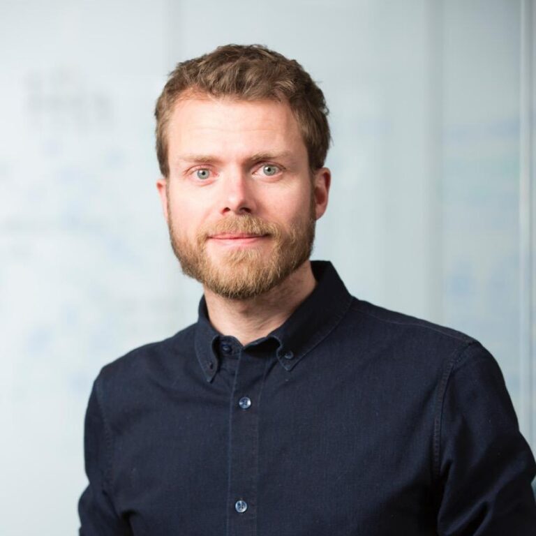 A man with light brown hair and a beard, wearing a dark button-up shirt, stands in front of a blurred whiteboard background, looking directly at the camera with a neutral expression.