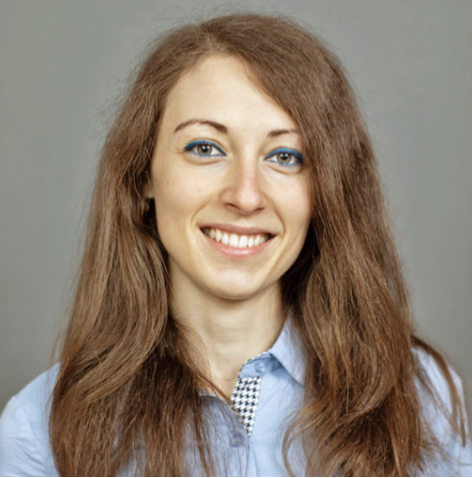 A young woman with long brown hair and blue eyeshadow smiles at the camera. She is wearing a light blue collared shirt and is posed in front of a plain gray background.