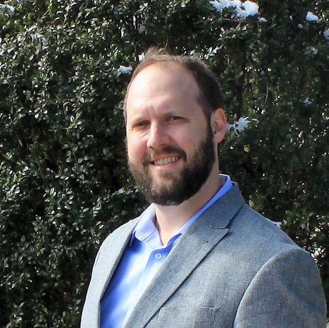 A man with a beard wearing a light gray blazer and blue shirt stands outside in front of green foliage with patches of snow.