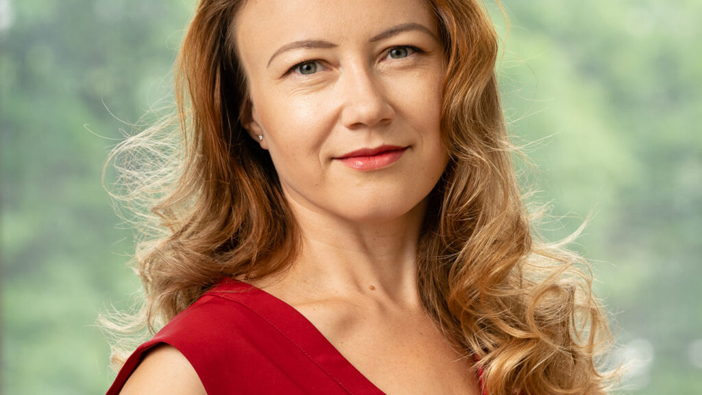 Woman with wavy, light brown hair wearing a sleeveless red top, smiling softly and standing in front of a blurred green outdoor background.