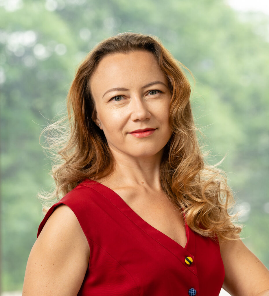 Woman with wavy, light brown hair wearing a sleeveless red top, smiling softly and standing in front of a blurred green outdoor background.