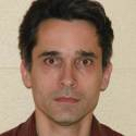 A man with short dark hair, fair skin, and brown eyes looks directly at the camera. He is wearing a maroon collared shirt and is posed against a plain, light-colored background.