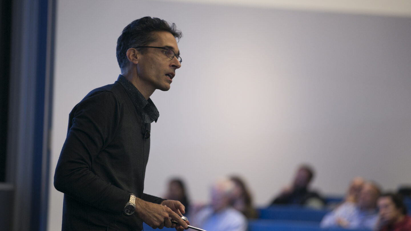 A man talking in front of a lecture hall while holding a metal stick in his hands