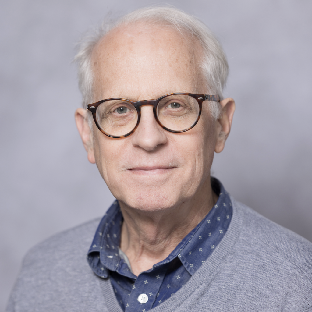 An older man with gray hair and glasses wears a blue collared shirt with a gray sweater. He is facing the camera and smiling softly. The background is plain and light gray.