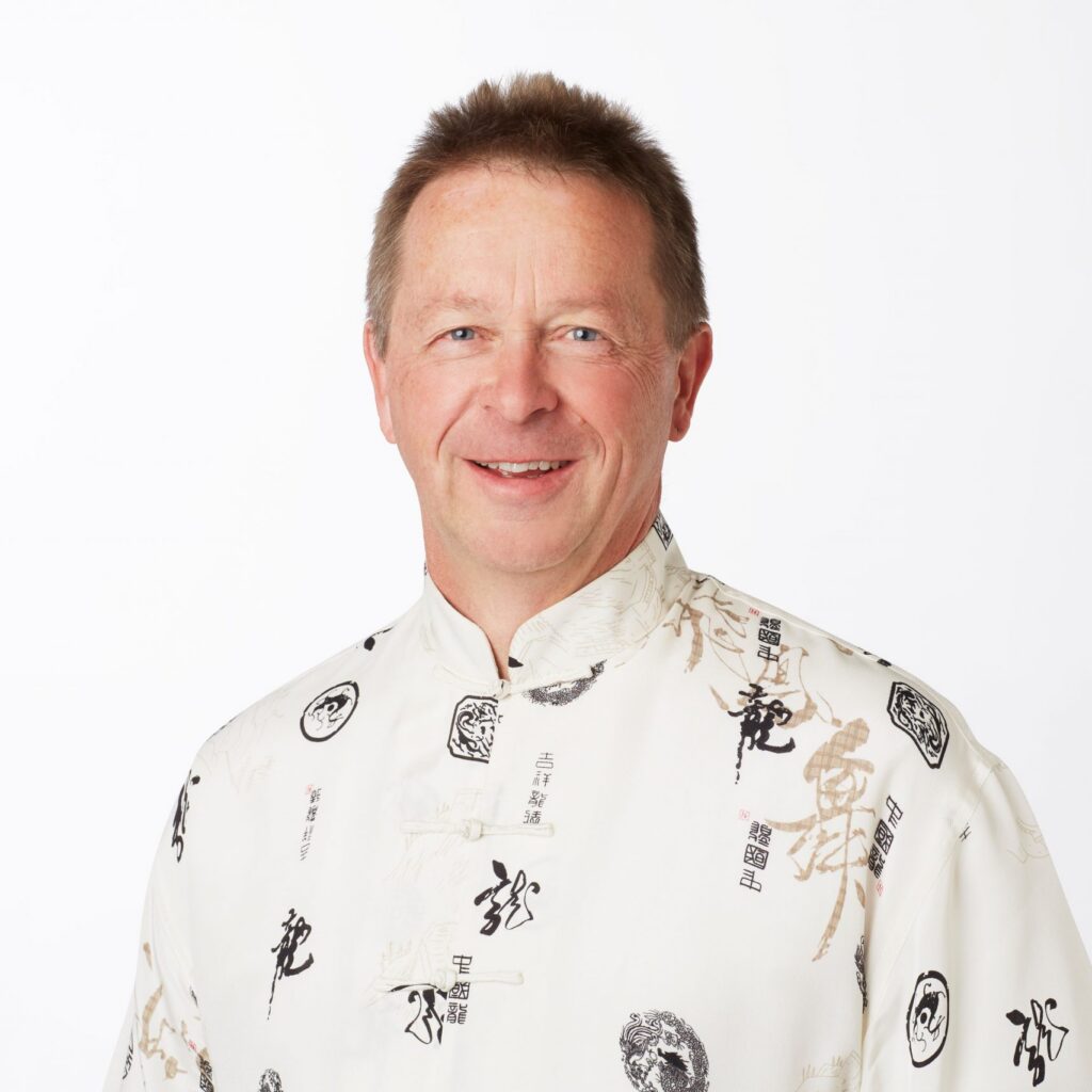 A smiling man wearing a white shirt with black and gold Chinese characters and symbols stands against a plain white background.